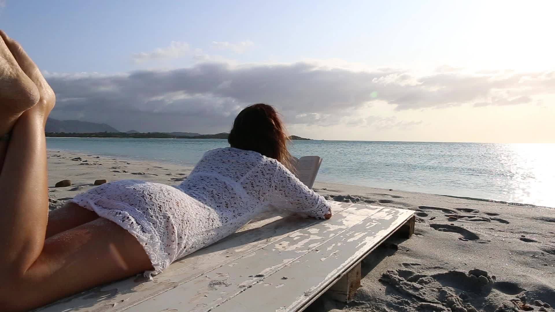 躺在海边的年轻女子穿着白色丝绸裙子、白色内衣和长发视频的预览图