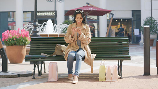 4k购物女性坐在长椅上休息使用手机视频的预览图