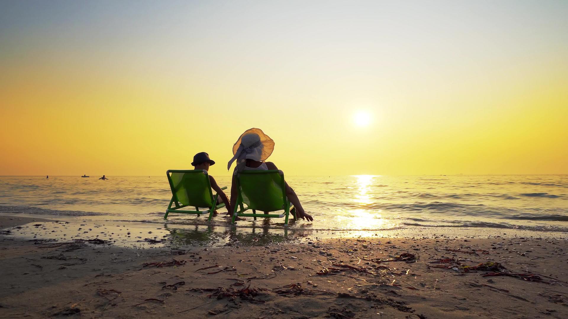 母亲和儿子戴着帽子坐在沙滩椅上看着夕阳旅行视频的预览图