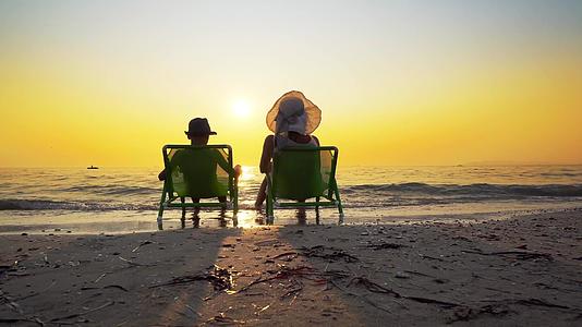 母亲和儿子戴着帽子坐在沙滩椅上看着海浪和日落视频的预览图