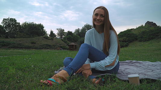 一个穿着绿衬衫的年轻女孩坐在公园的草坪上喝着酒微笑着视频的预览图