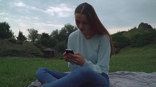 一个年轻女孩坐在公园草坪上看手机视频的预览图