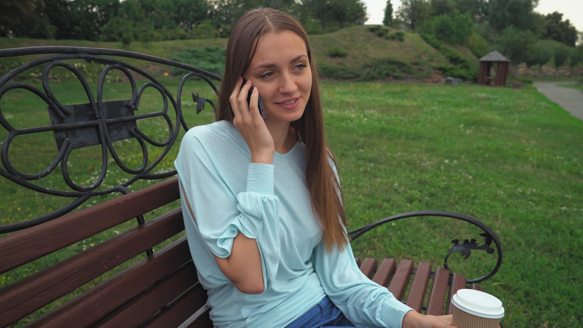 一个年轻女孩坐在公园长椅上喝酒在电话里微笑着说话视频的预览图