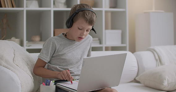 小男孩坐在沙发上用笔记本电脑在纸上画画视频的预览图