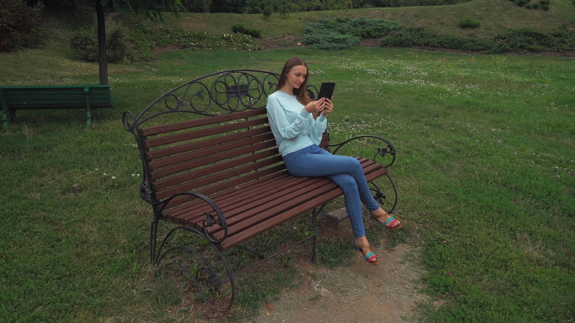 留着长发的年轻女孩坐在公园长椅上拿着平板电脑微笑着视频的预览图