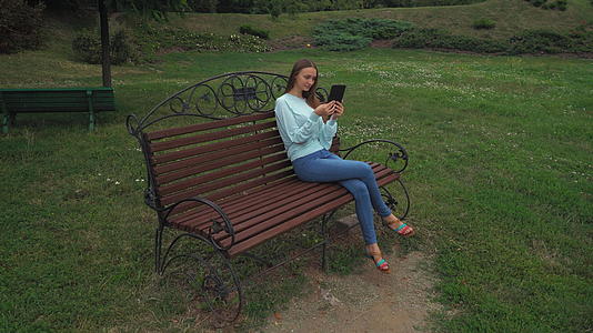 留着长发的年轻女孩坐在公园长椅上 拿着平板电脑 微笑着视频的预览图