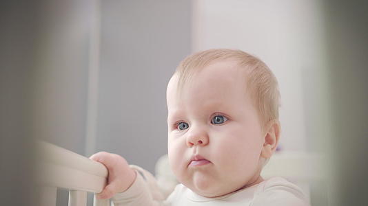 婴儿站在床上用蓝眼睛看着可爱的婴儿脸孔视频的预览图