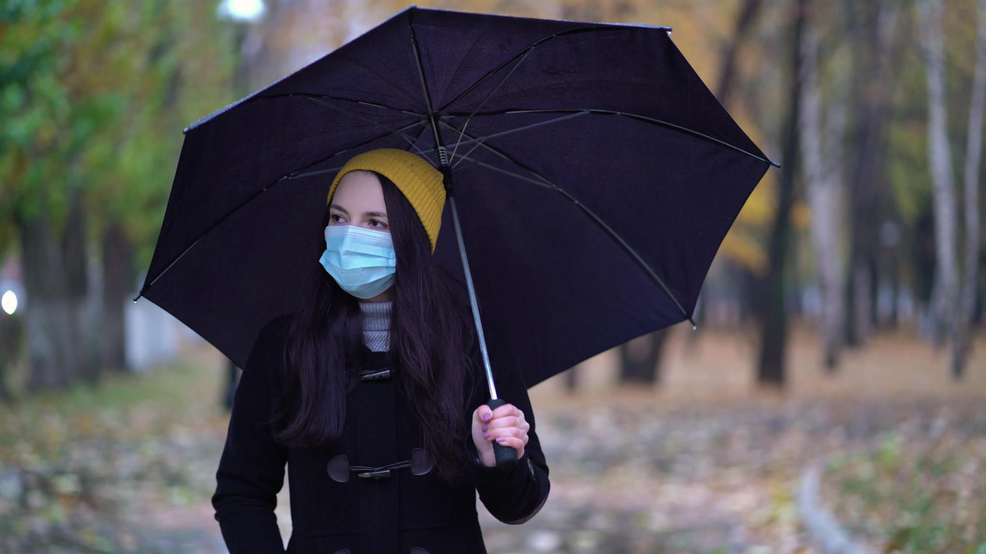 一个戴着防护面具的年轻女子在雨伞下在公园里散步视频的预览图