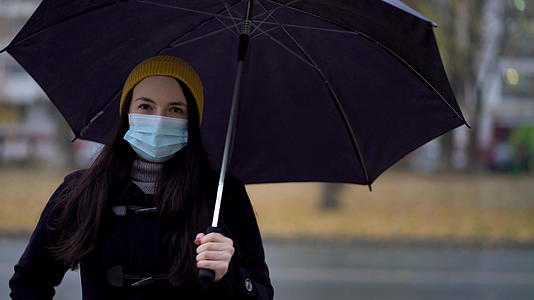 一个戴着防护面具的年轻女子在雨伞下的公园里散步视频的预览图