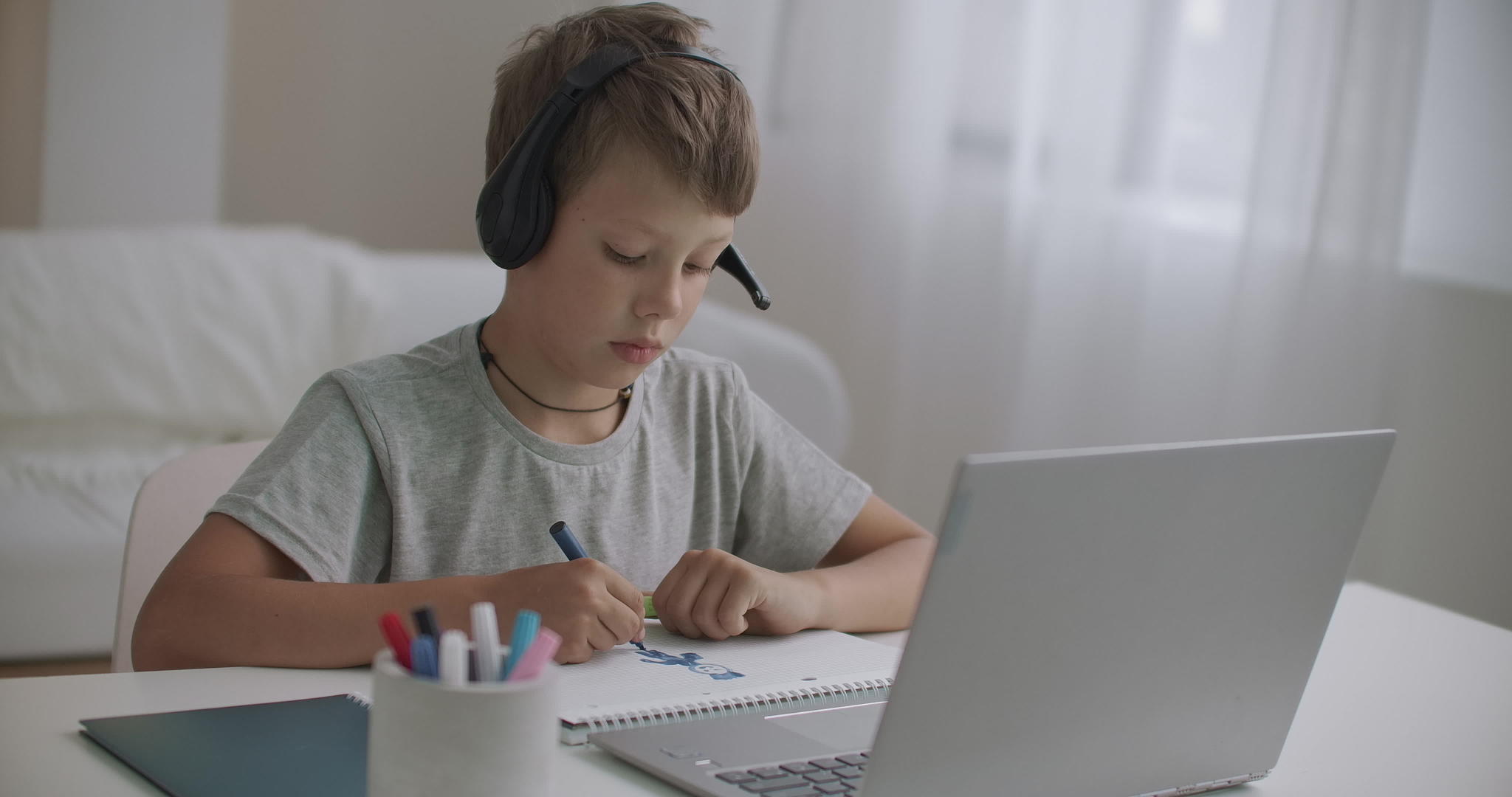 在大流行中在家上学的小男孩正在用耳机听老师用笔记本电脑讲课视频的预览图