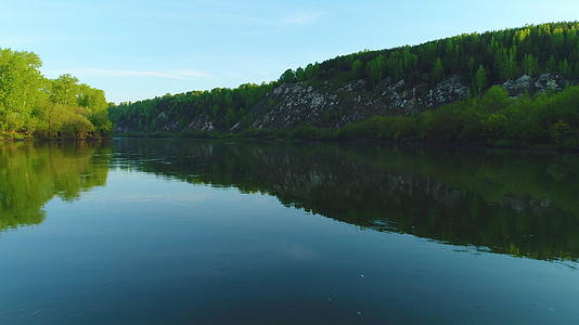 在阳光明媚的白天在河边的低角飞过河边有绿树的岩石和蓝天视频的预览图
