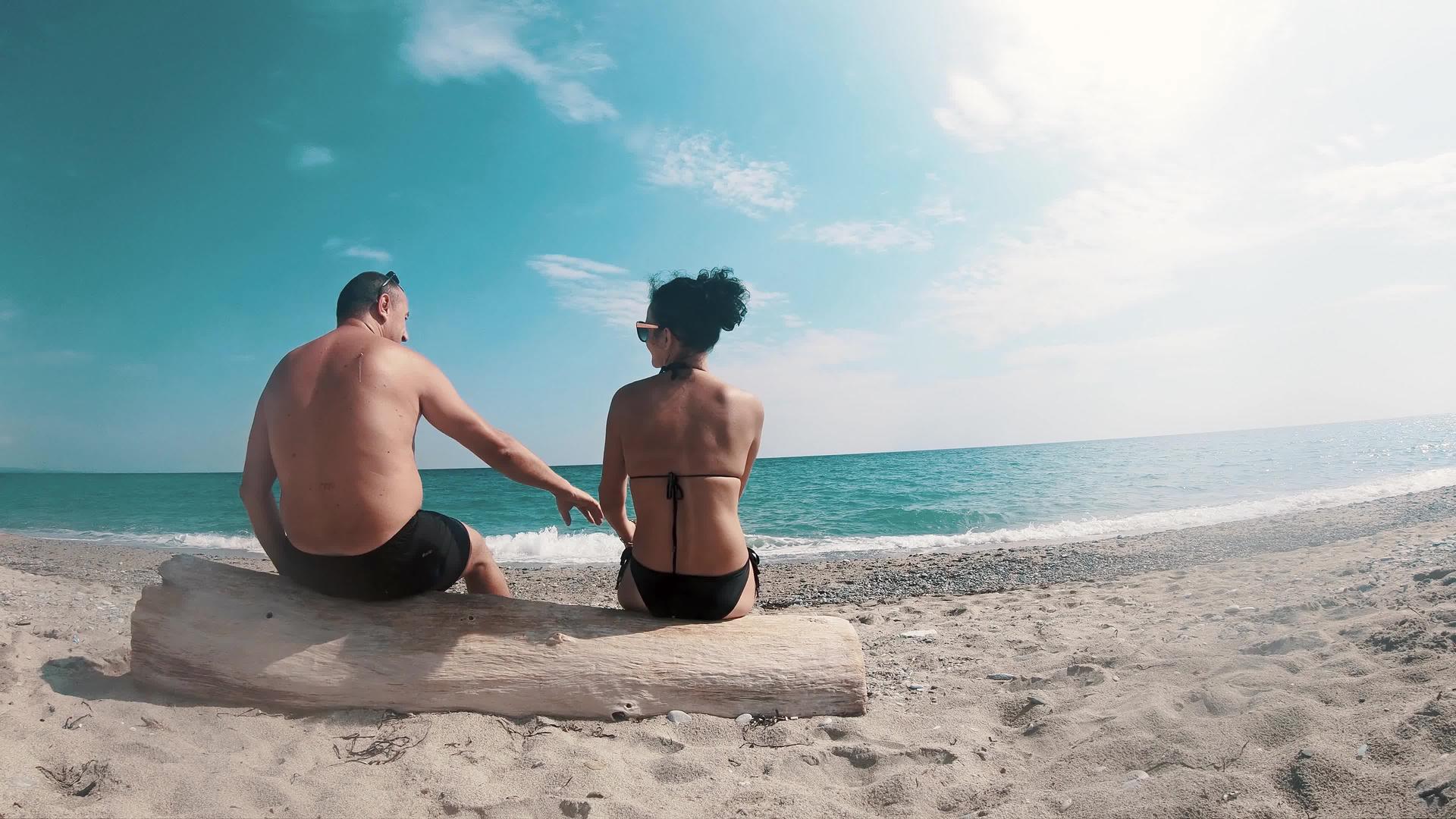 恋爱中的恋人握手坐在空荡荡的海滩树干上视频的预览图