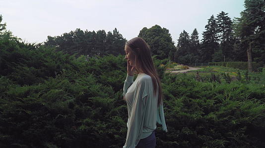 年轻女子沿着公园的灌木丛走在电话里视频的预览图