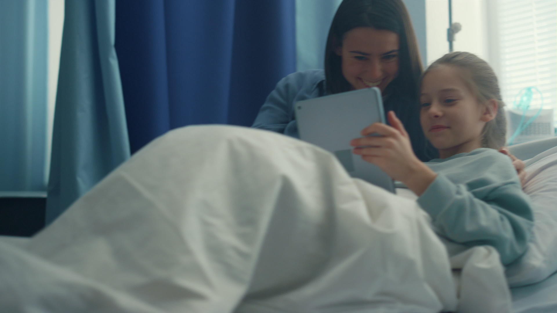 微笑的母亲看望女儿在医院病房玩平板电脑视频的预览图