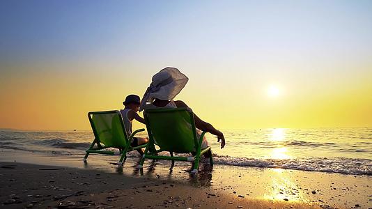 母亲和儿子坐在沙滩甲板椅上面对日落电影制片人Stadicam视频的预览图