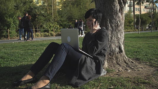 女人在笔记本电脑上打字在公园的树下用智能手机说话视频的预览图