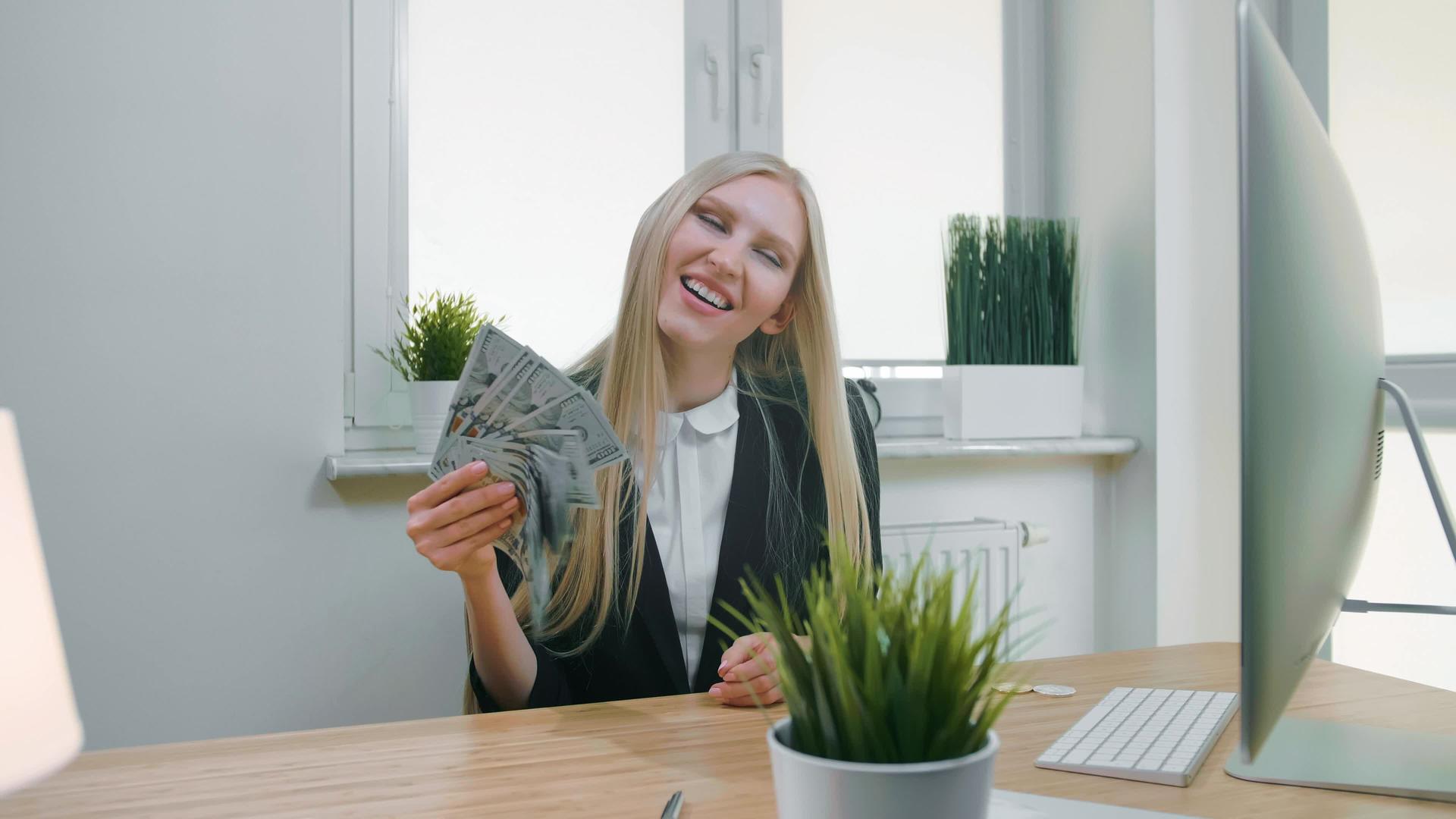 美丽快乐的金发女人坐在办公室里微笑着拿着一堆现金视频的预览图