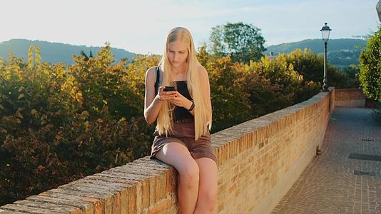 坐在阳光明媚的街道上的年轻女性使用智能手机视频的预览图