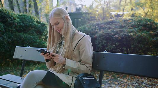 金发女孩在公园的户外长椅上使用手机视频的预览图