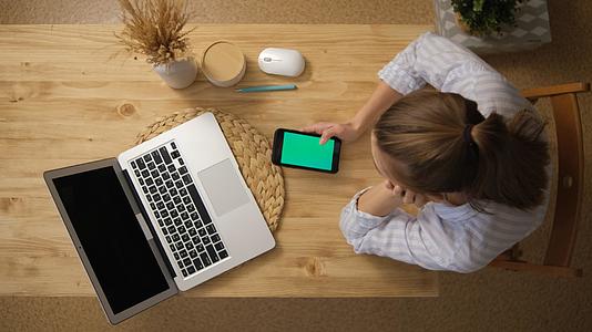 女孩用绿屏看着手机对面是一台带有绿色屏幕的开放式笔记本电脑视频的预览图