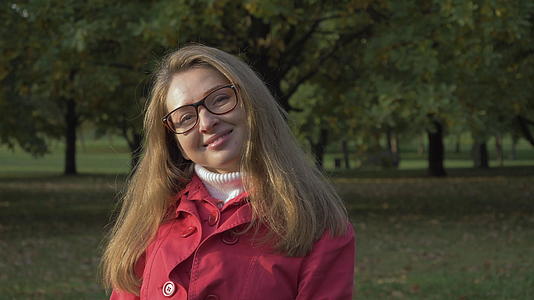 戴着眼镜和红色夹克的绿色公园肖像中年轻微笑的女士视频的预览图