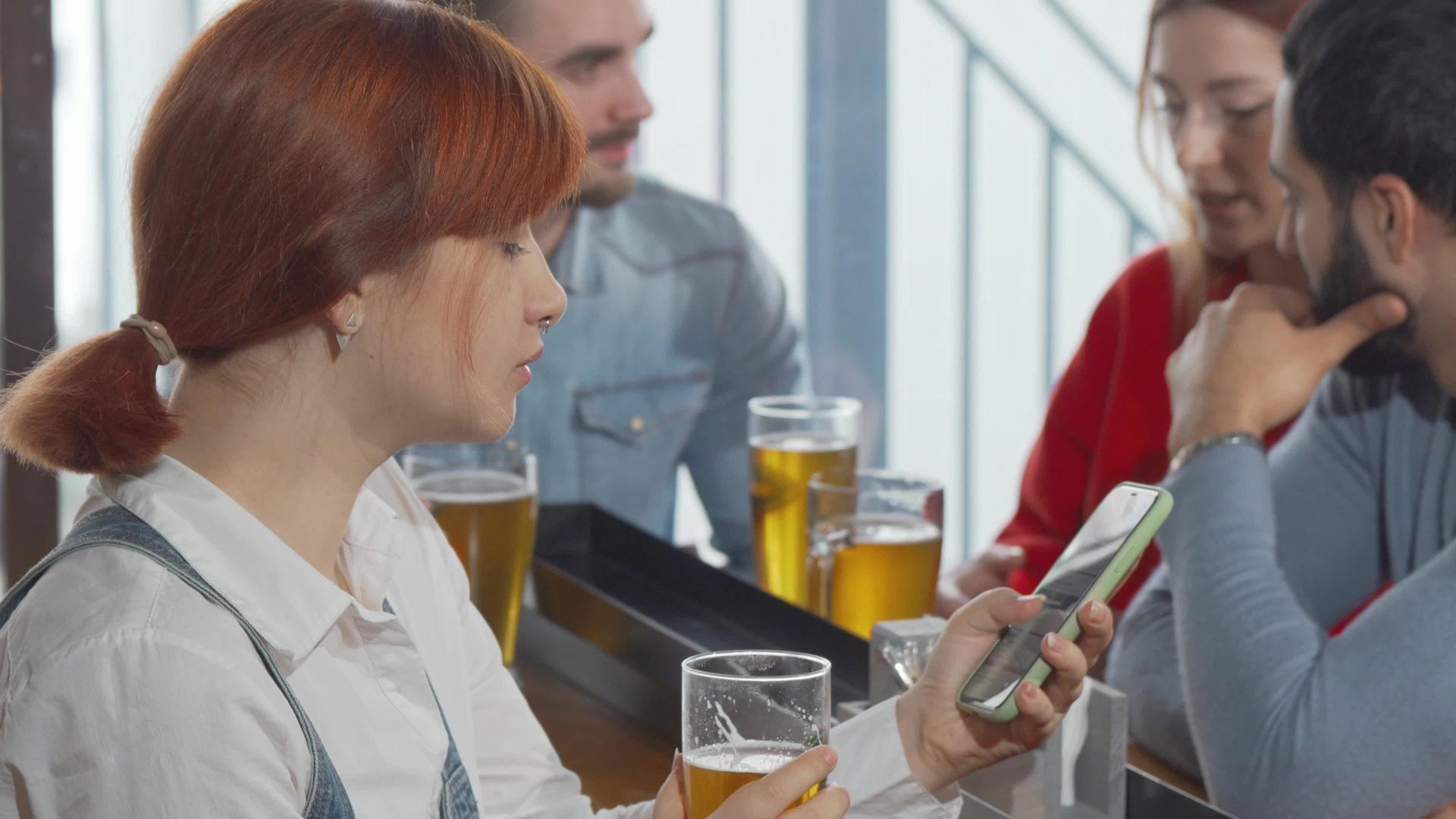 年轻女性在酒吧喝啤酒时使用智能手机和啤酒视频的预览图