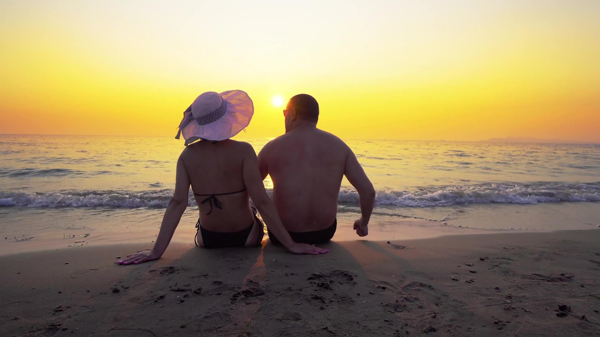一对戴着帽子的浪漫情侣男女坐在海看着日落视频的预览图