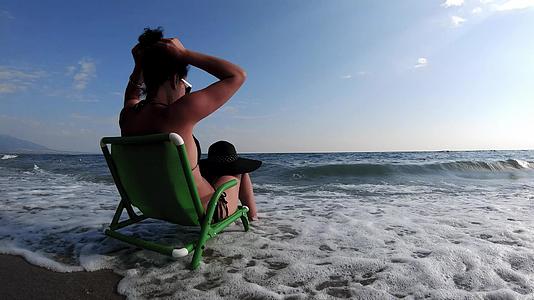 穿着比基尼的漂亮年轻美女戴着帽子坐在沙滩上的椅子上视频的预览图