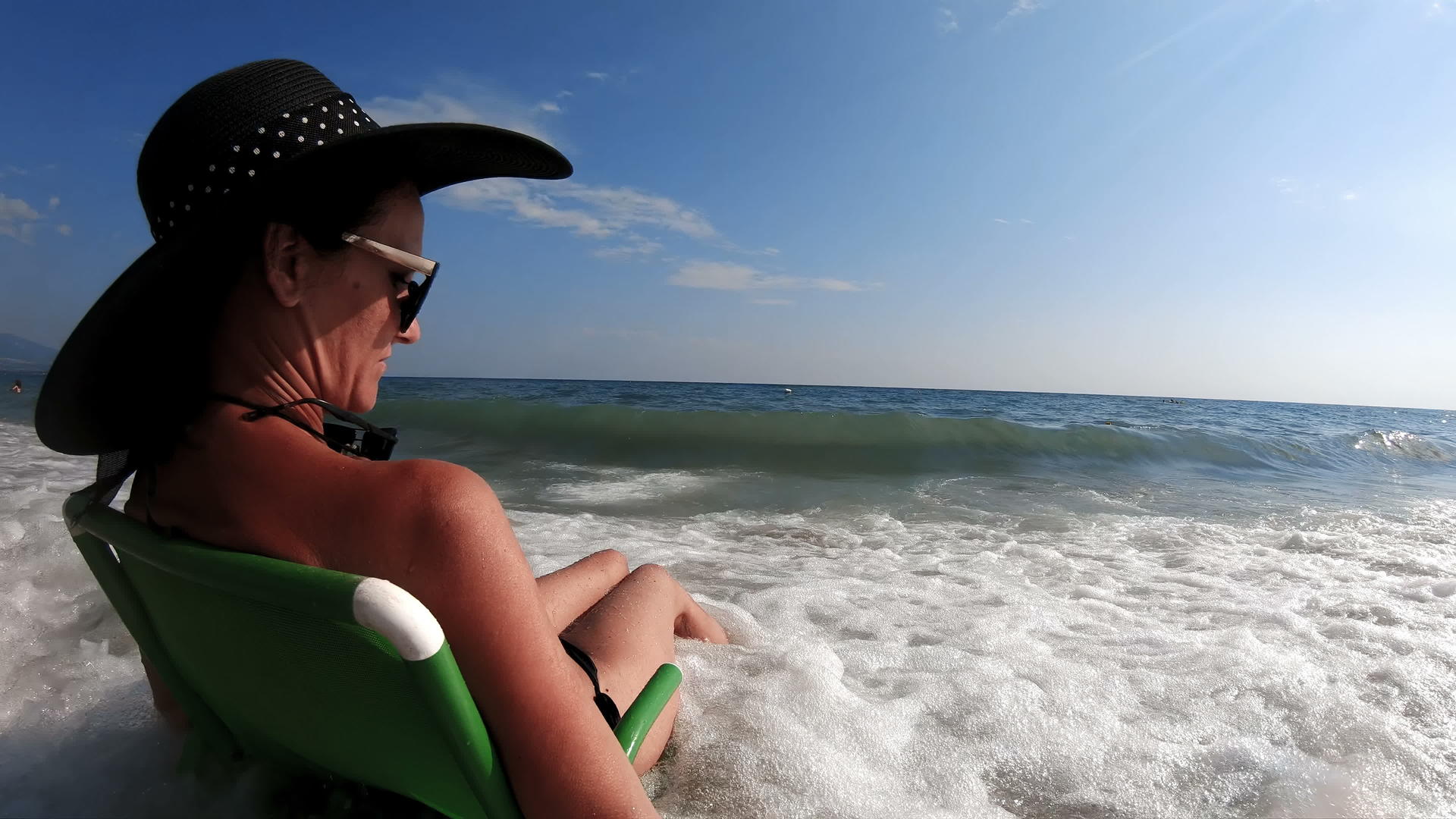 迷人的女人坐在海滩上戴着草帽享受着海浪在岸边坠落的乐趣视频的预览图