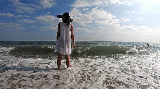 一个穿着草帽和白色连衣裙的女人站在浅海享受着海浪的味道视频的预览图