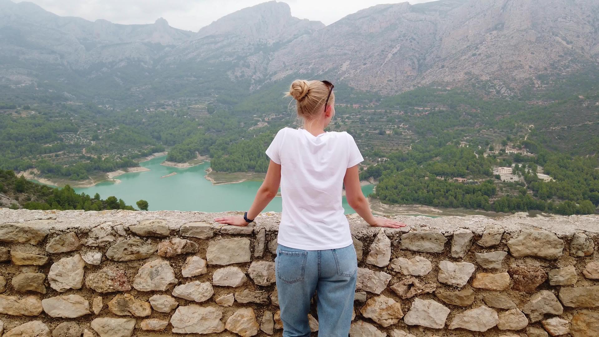 一个年轻的女孩从一座古老的城堡里看着一个美丽的风景蓝湖和山峰视频的预览图