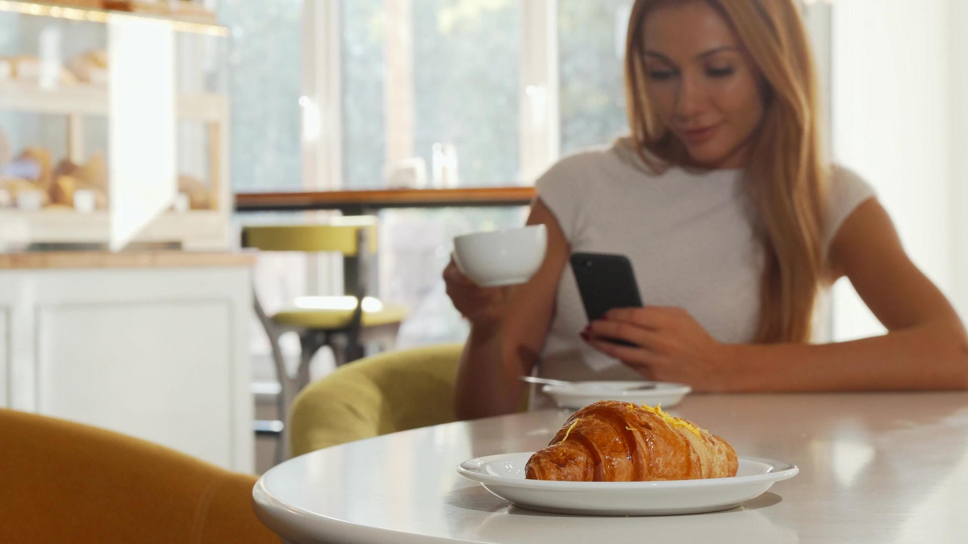 在咖啡店使用智能手机的女性选择性地关注美味的羊角面包视频的预览图
