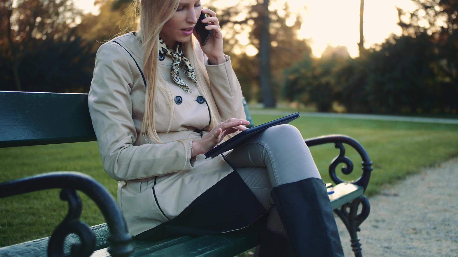 一个漂亮的女孩坐在公园长椅上平板电脑和手机视频的预览图