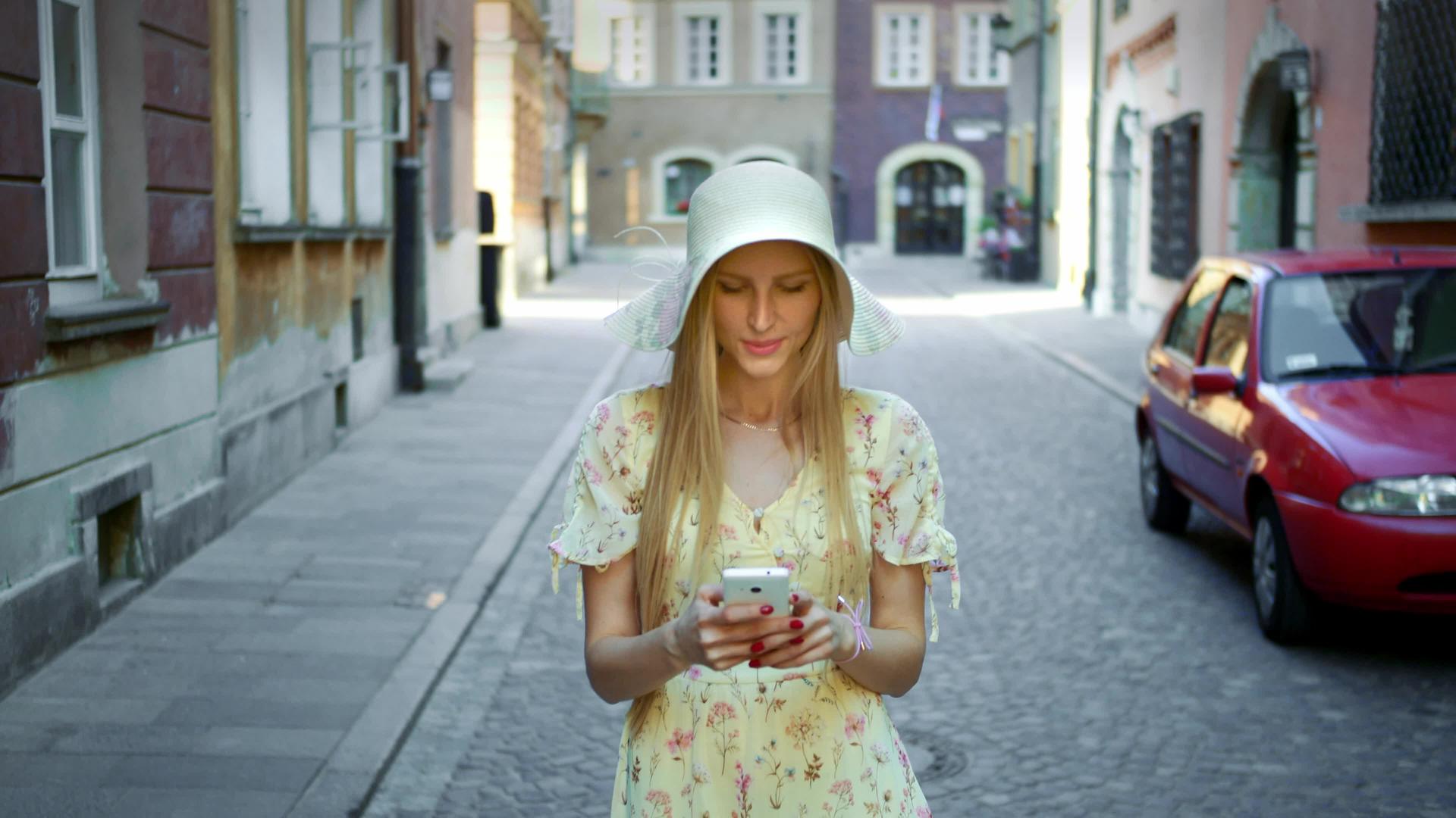 一位戴着白帽子的年轻女士走在老城街上用智能手机给她发短信视频的预览图