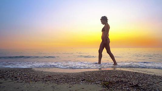 日落时在海滩上散步的合身女人在海浪上喷水视频的预览图