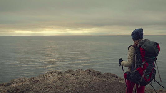 背着背包和登山杖的年轻女子走到海边的岩石边缘视频的预览图