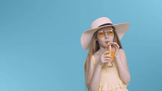 穿着黄色眼镜和帽子的年轻女孩在蓝屏上喝鸡尾酒视频的预览图