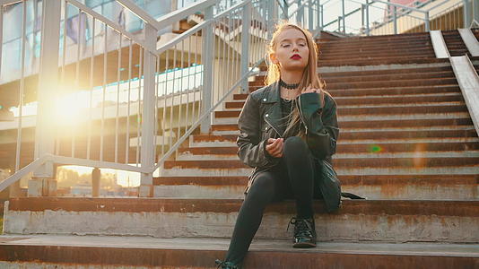 一个孤独的黑色皮夹克女孩坐在阳光背景的楼梯上视频的预览图