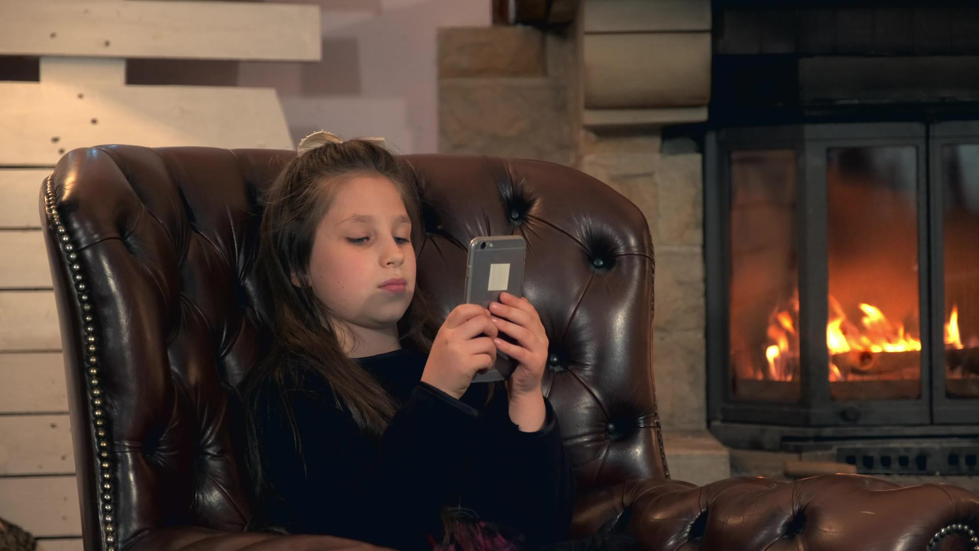 一个漂亮的小女孩在壁炉附近的智能手机上玩游戏视频的预览图