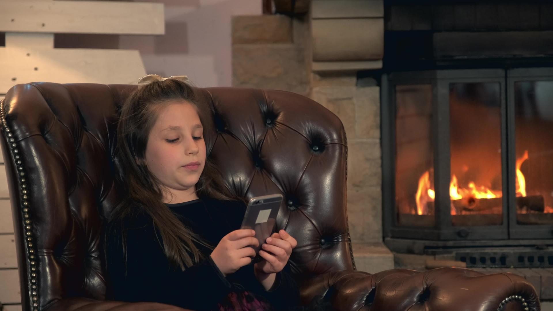 小女孩在壁炉附近使用智能手机的闲暇时间视频的预览图