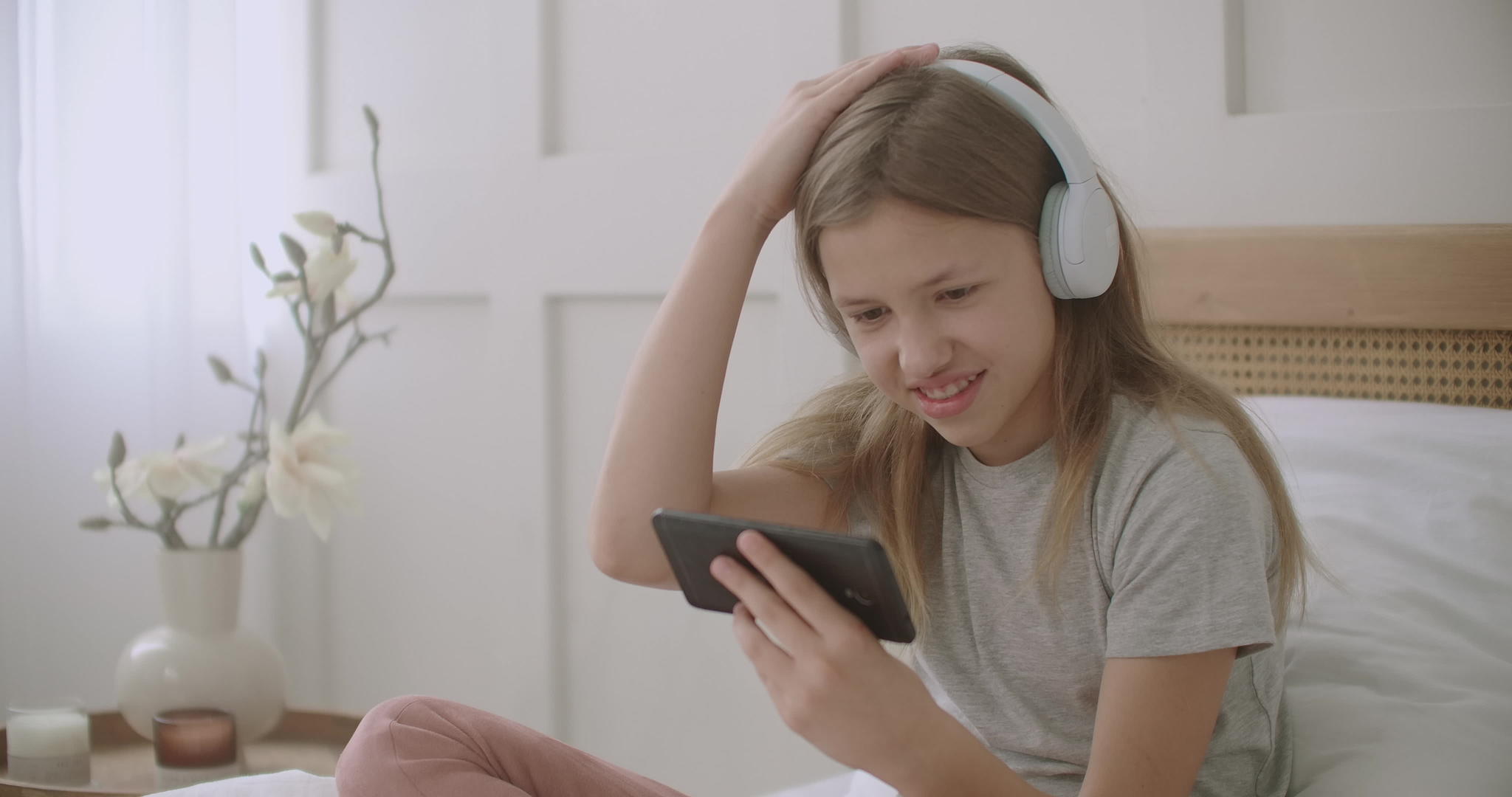 小女孩正在用智能手机和无线耳机坐在房间的床上玩游戏视频的预览图