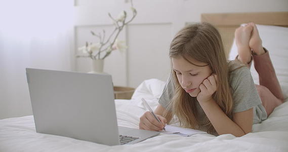 小女孩正躺在房间里的笔记本电脑前在书里画画和写字视频的预览图