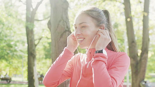 在公园里跑步时穿着粉色运动服的健身女孩用耳机听音乐和音乐视频的预览图