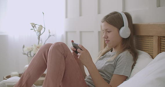 女学生呆在家里玩手机游戏躺在床上用耳机听音乐视频的预览图