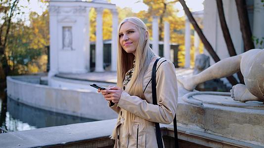 秋天公园里穿着米花大衣的金发美女看着她的智能手机视频的预览图