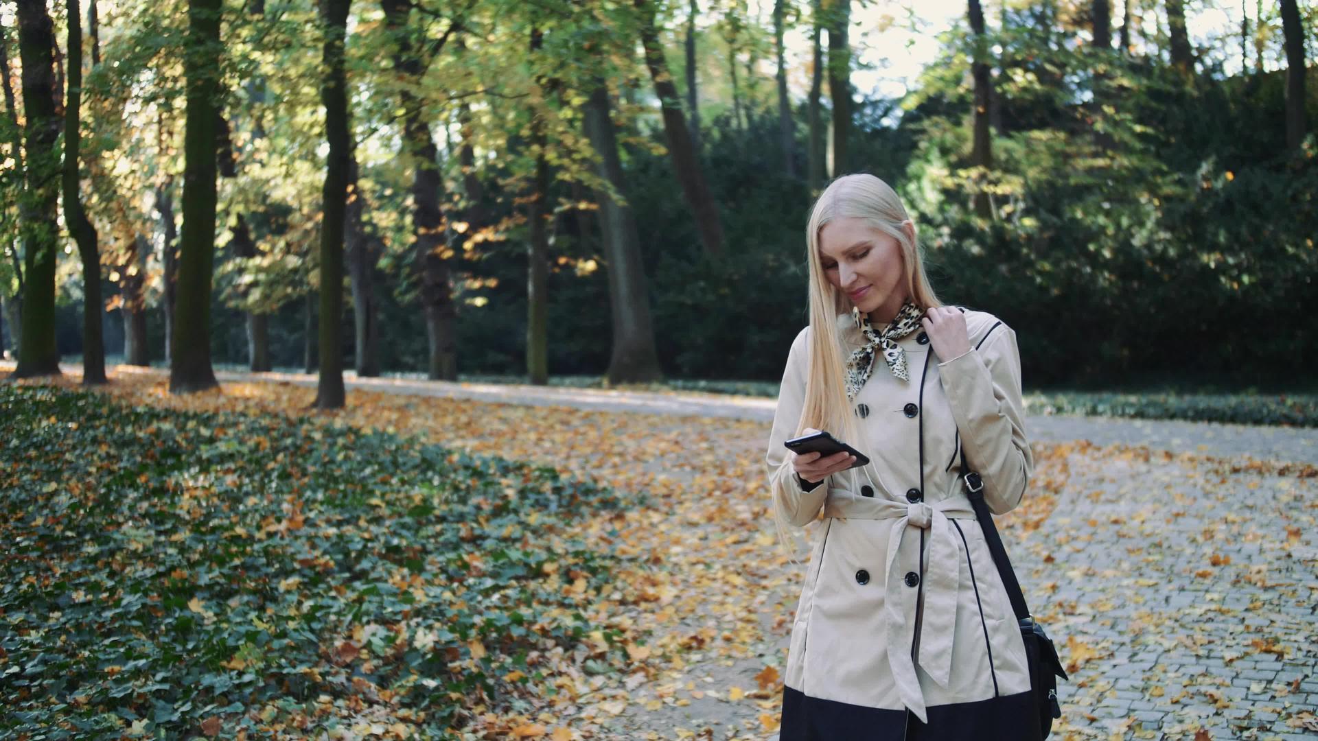 秋天我在公园里散步看着金发女的智能手机和打字视频的预览图