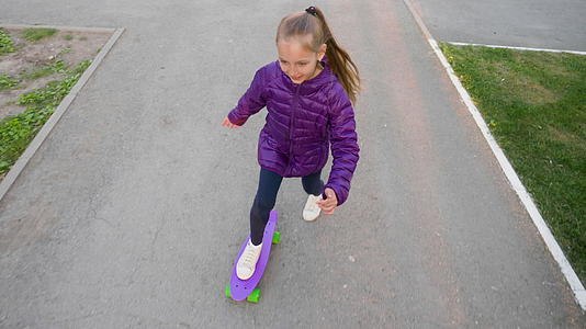 快乐的女孩穿着夹克在公园街的紫色滑板上滚动视频的预览图
