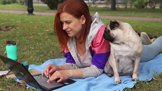 女孩躺在笔记本电脑上打字她的狗在草坪上视频的预览图