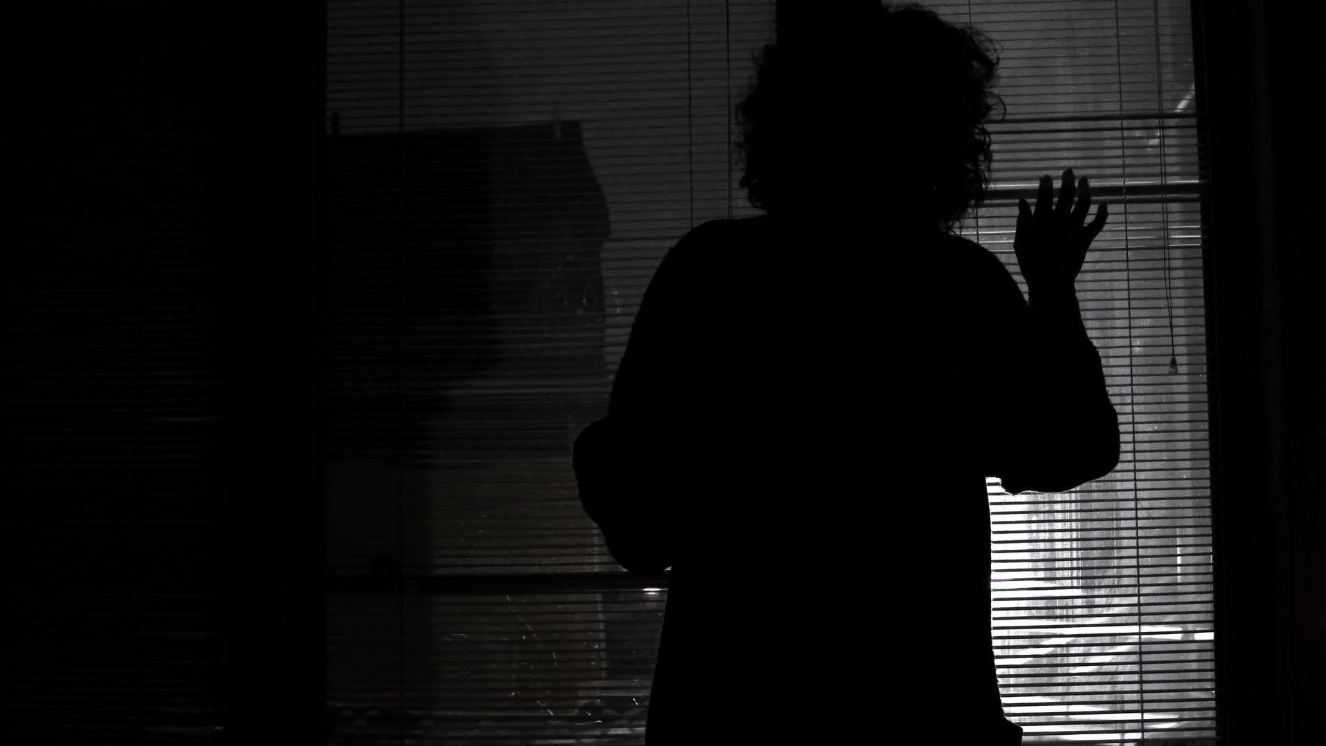 在黑暗的夜晚在户外的灯光下惊恐的剪影女人透过百叶窗向外看着视频的预览图