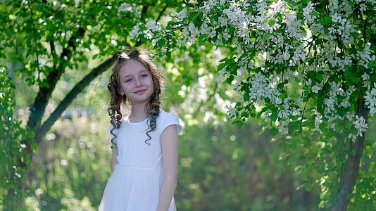 微笑的卷发女孩肖像在阳光明媚的花园里盛开的果树背景下视频的预览图